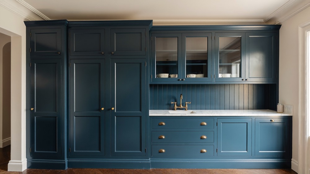 A wall of deep blue painted cabinetry with tall panelled doors, glazed uppers, brass cup pulls and a small marble sink