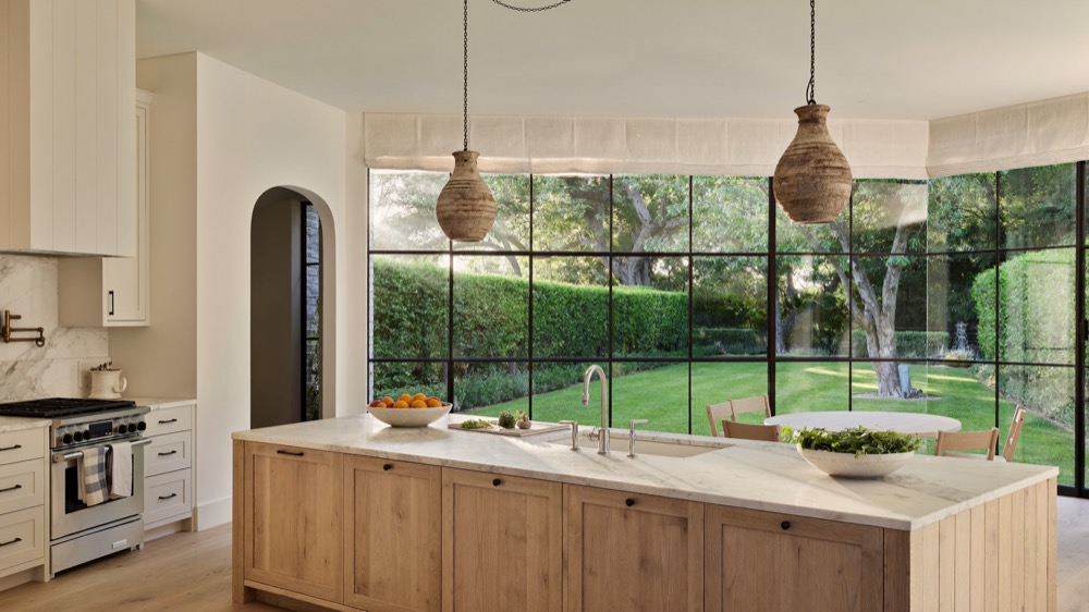 An oak island with a marble top in front of tall windows looking onto a lawn and hedges