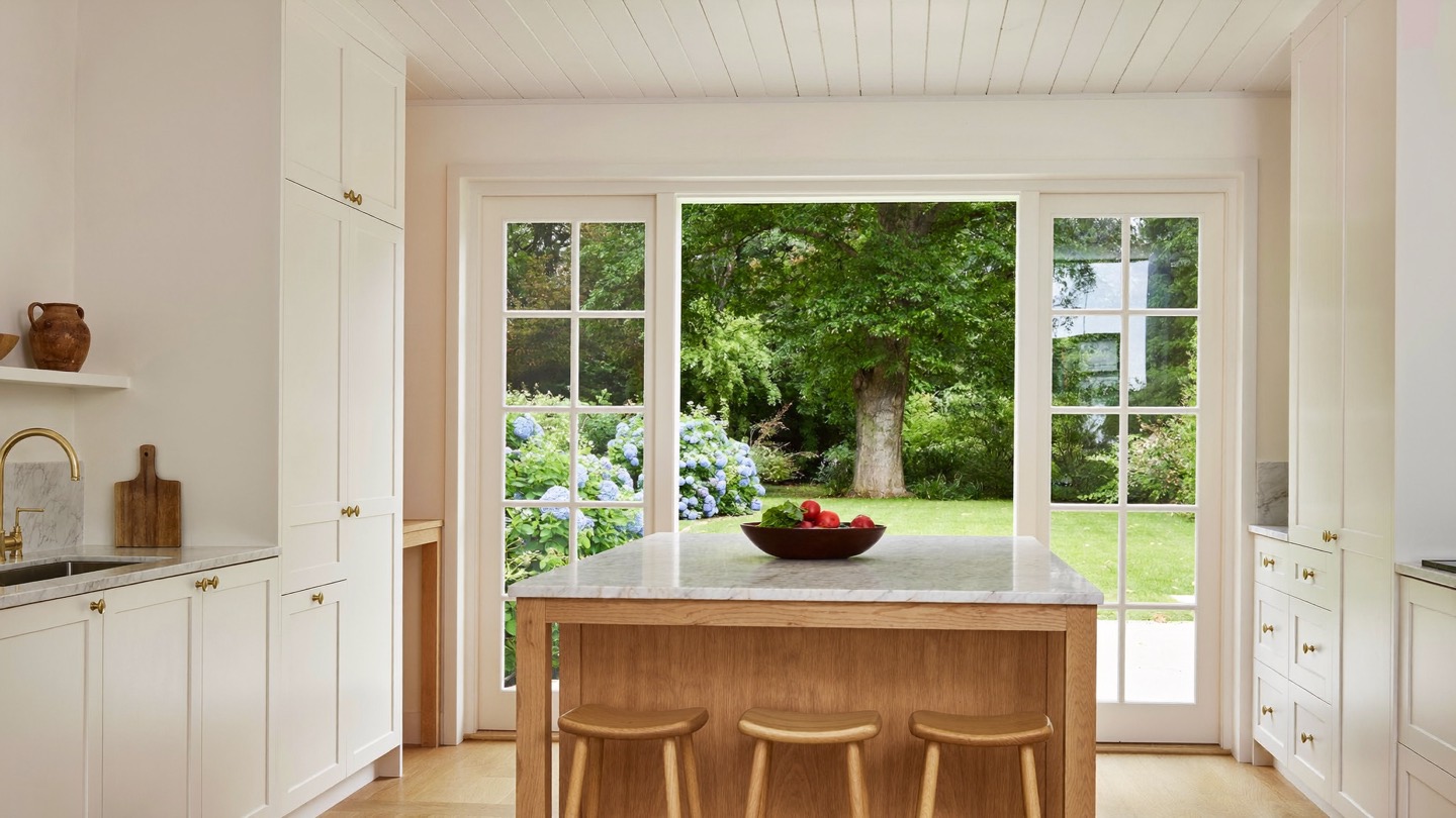 A white painted kitchen with an oak island, French doors open to a lawn and blue hydrangeas