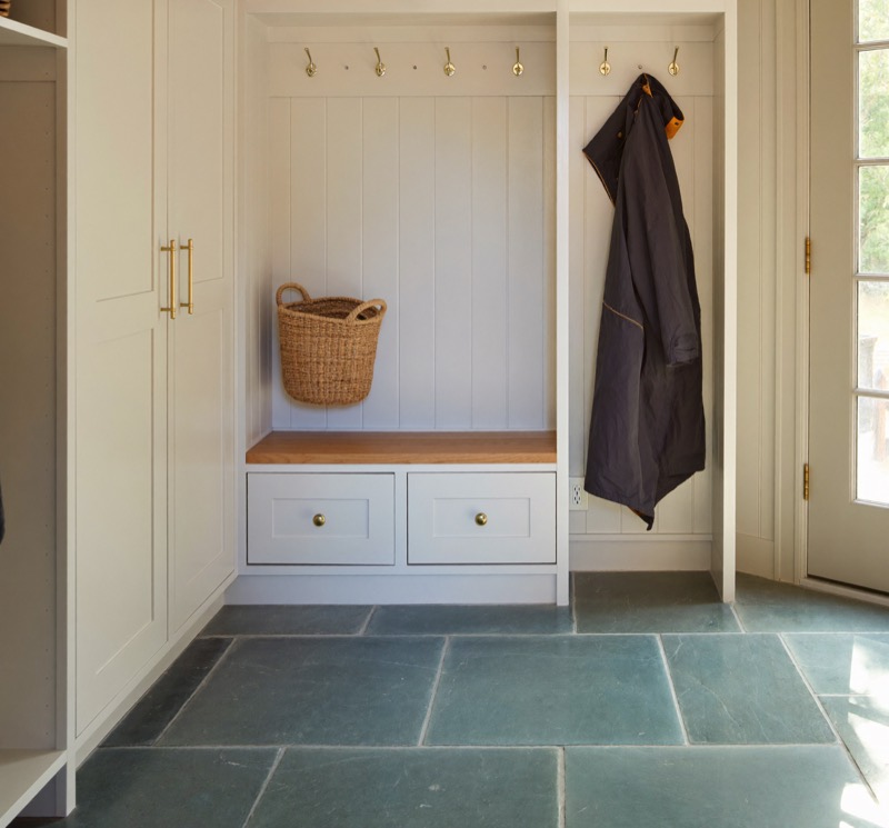 A painted mudroom built-in with brass hooks, an oak bench and drawers over a bluestone floor