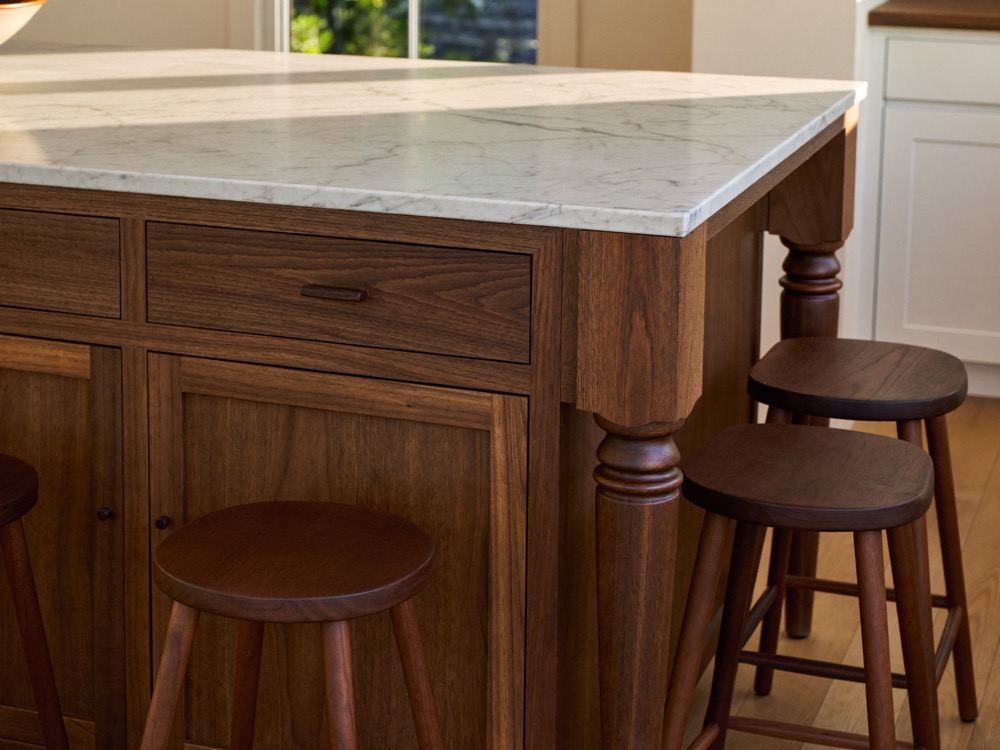 Corner of a walnut kitchen island with a turned leg, a white marble top and walnut stools